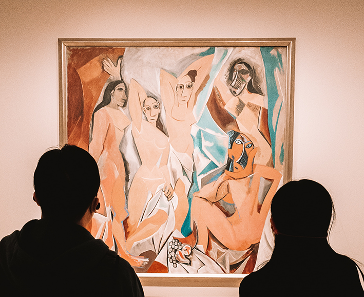 Zwei Personen stehen vor Picassos Les Demoiselles d'Avignon, einem kubistischen Gemälde mit fünf abstrakten Figuren, und diskutieren über Kunstschätzung und Kunstbewertung, während sie die kühnen Formen und gedämpften Farben an der Museumswand bewundern.