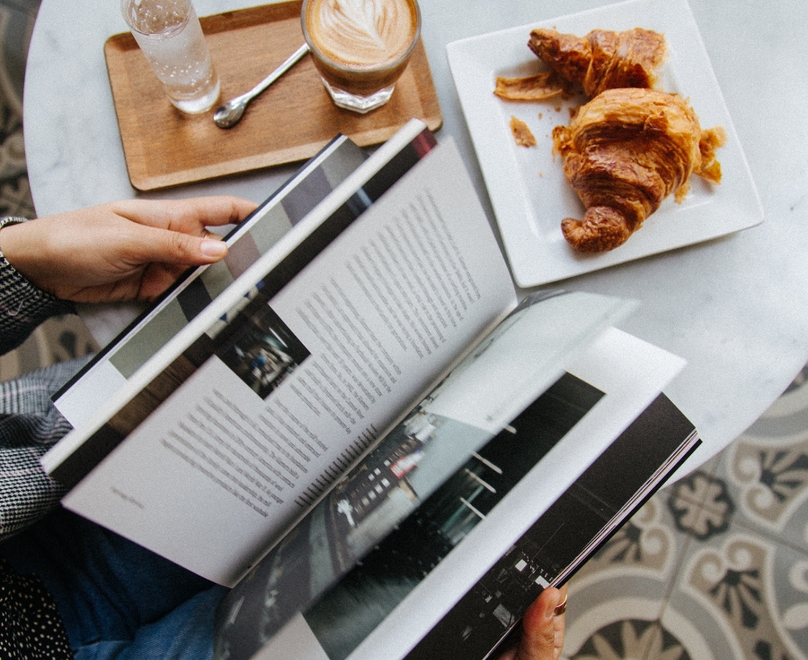 Eine Person liest ein aufgeschlagenes Buch an einem Tisch mit einem Milchkaffee, einem Glas Wasser auf einem Holztablett und einem Teller mit einem Croissant. Der gemusterte Tisch deutet auf eine Wertschätzung der Kunstsammlung in alltäglichen Momenten hin.